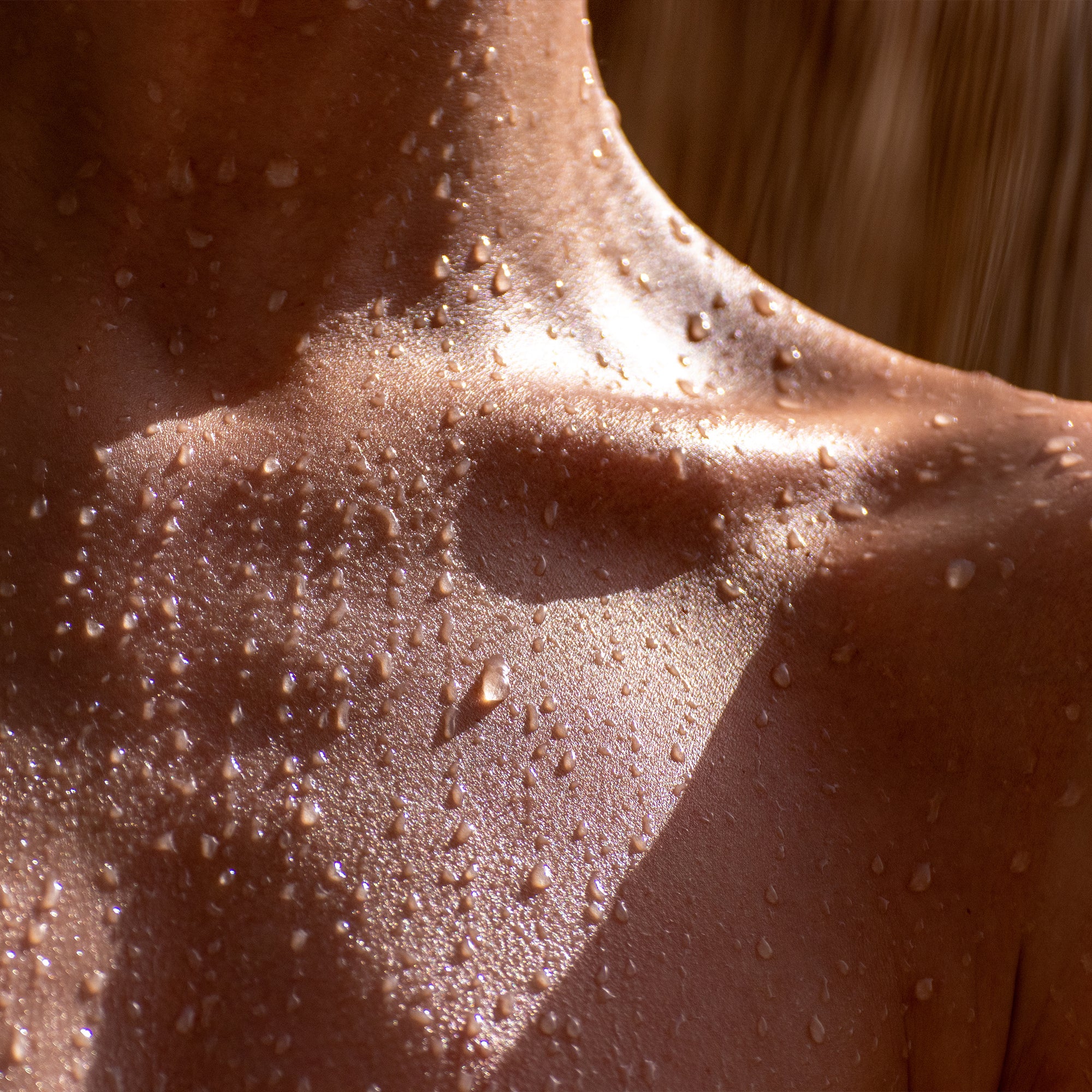 Detailaufnahme eines nackten oberen Brustbereichs mit glänzender, feuchter Haut, bedeckt von Wassertropfen. Das Licht betont die natürliche Textur und die gesunde Ausstrahlung der Haut.