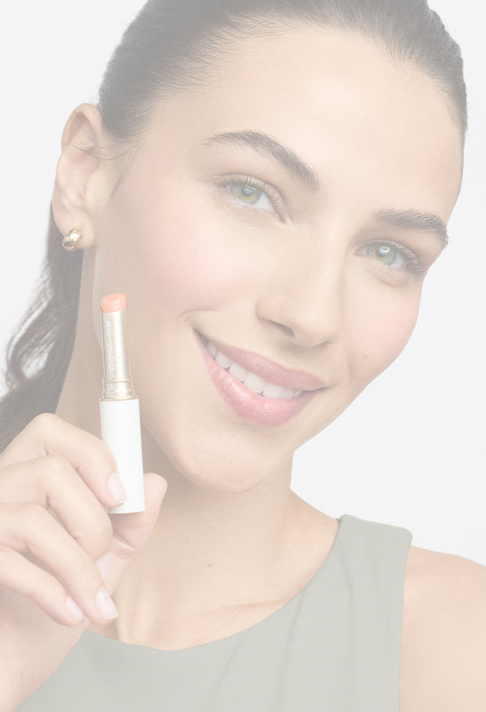 Frau mit glatter, strahlender Haut hält einen Lippenpflegestift von jane iredale in der Hand und lächelt freundlich in die Kamera vor grauem Hintergrund.