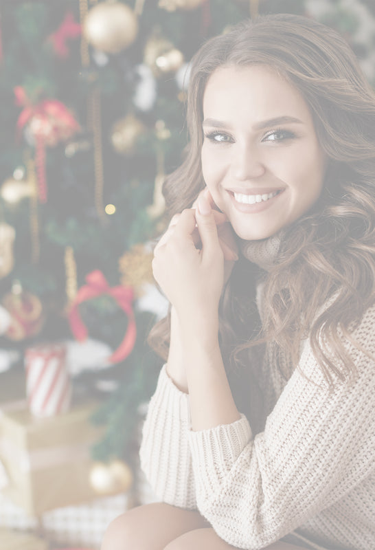 Junge Frau mit langen, gewellten Haaren sitzt lächelnd vor einem festlich geschmückten Weihnachtsbaum mit goldenen und roten Kugeln sowie Geschenken im Hintergrund.