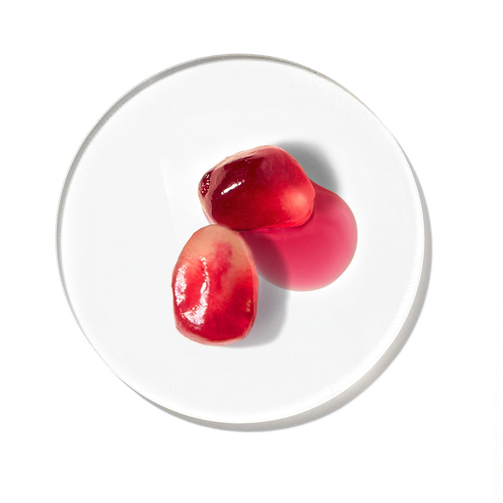 Zwei rote Granatapfelkerne mit einem rosafarbenen Öltropfen auf einer runden, transparenten Glasschale vor weißem Hintergrund.