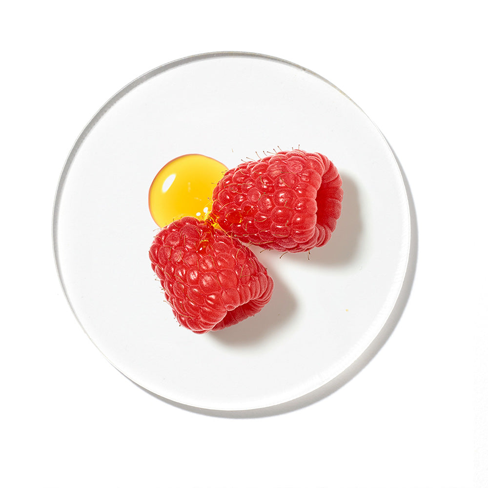 Zwei frische rote Himbeeren neben einem goldgelben Öltropfen auf einer runden, transparenten Glasschale vor weißem Hintergrund.