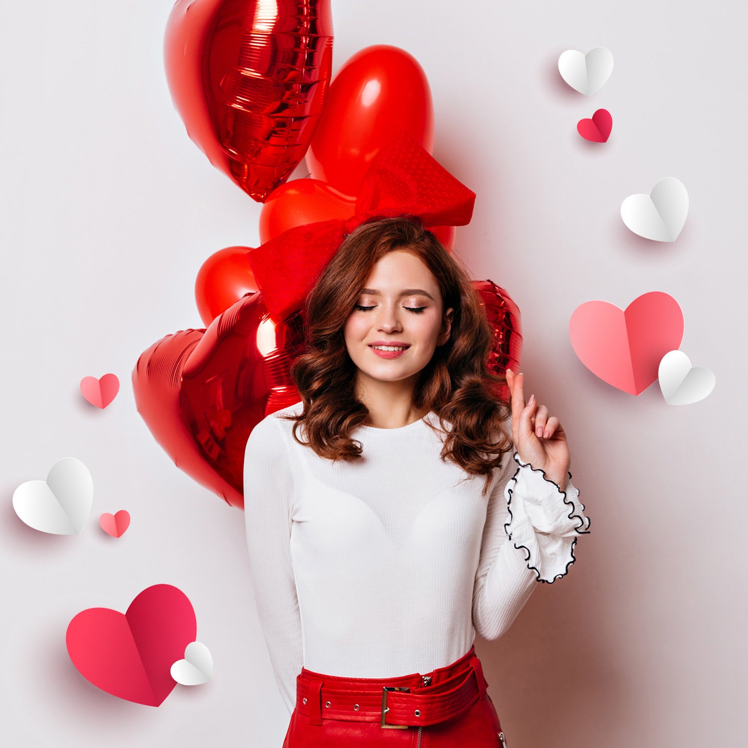 Junge Frau mit schulterlangen lockigen Haaren trägt ein weißes Oberteil und einen roten Rock. Sie steht vor mehreren roten Herzballons und hält eine Hand seitlich erhoben. Der Hintergrund ist hell, dekoriert mit roten, rosafarbenen und weißen Papierherzen.