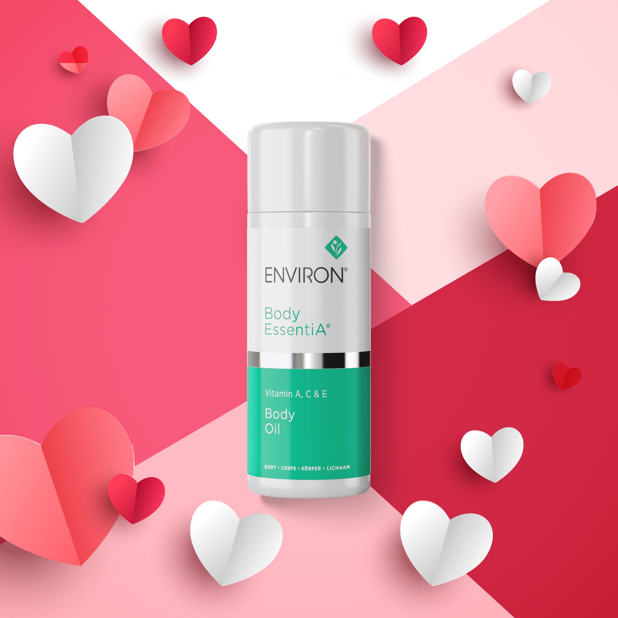 Zylindrische Flasche „Environ Body EssentiA – Body Oil“ mit weißem und türkisfarbenem Etikett. Die Flasche steht auf einem geometrisch geteilten rosa-roten Hintergrund, dekoriert mit Papierherzen in Rot, Rosa und Weiß.