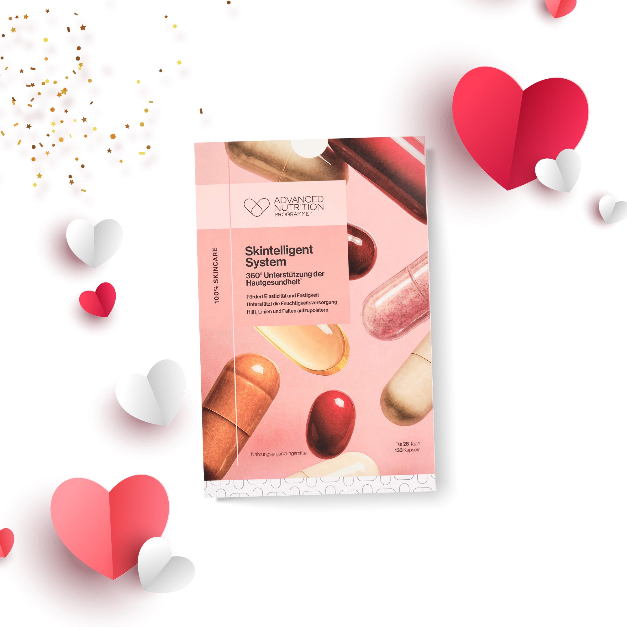 Produktverpackung „Skintelligent System“ mit Abbildungen verschiedener farbiger Kapseln auf dem Design. Die Packung befindet sich auf einem weiß-rosa Hintergrund und ist von Papierherzen in Rot, Rosa und Weiß sowie goldenen Konfettielementen umgeben.