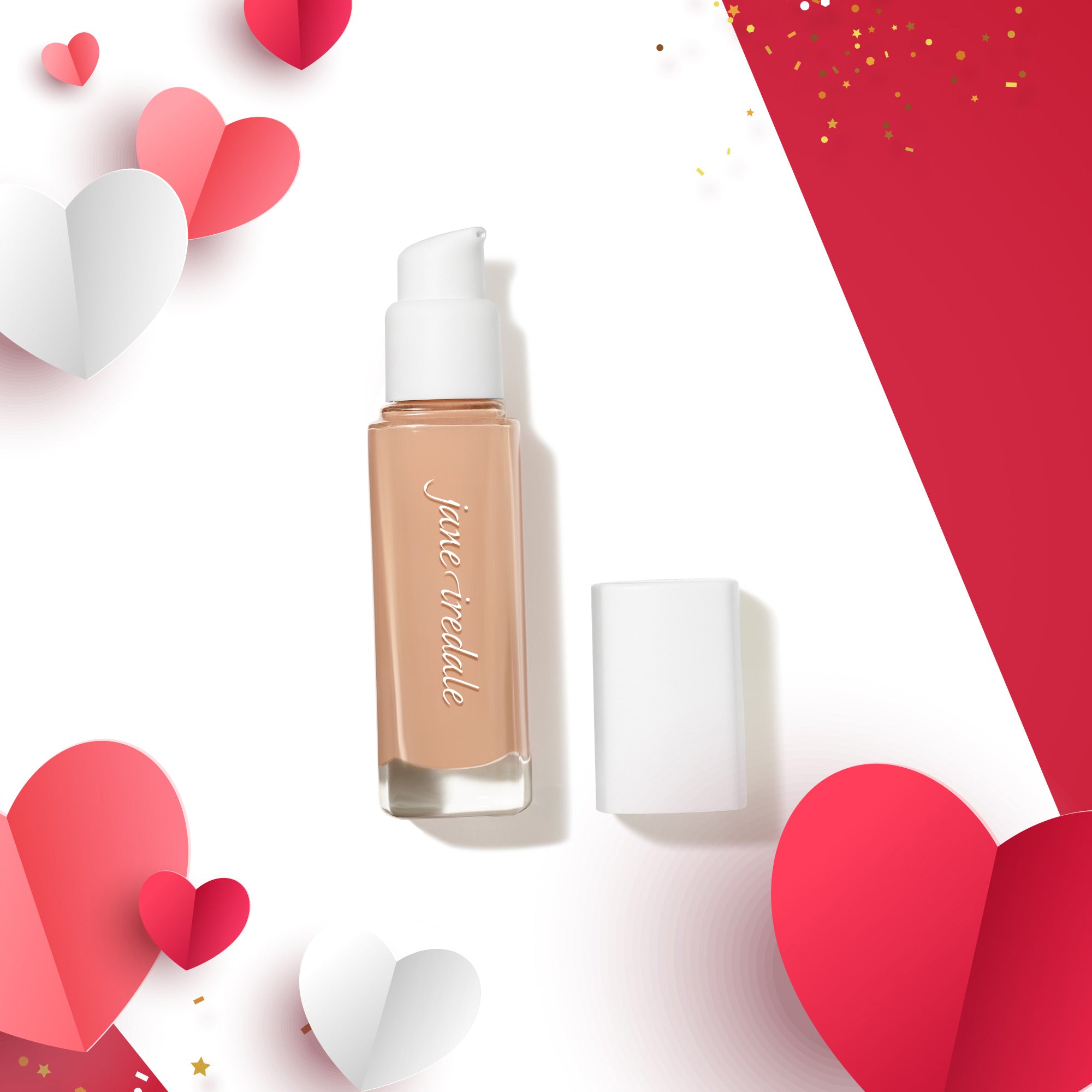 Rechteckige Pumpspenderflasche mit beiger Foundation und weißem Pumpverschluss, Deckel daneben liegend. Die Flasche befindet sich auf einem rot-weißen Hintergrund mit dekorativen Papierherzen in Rosa, Rot und Weiß sowie goldenem Konfetti.