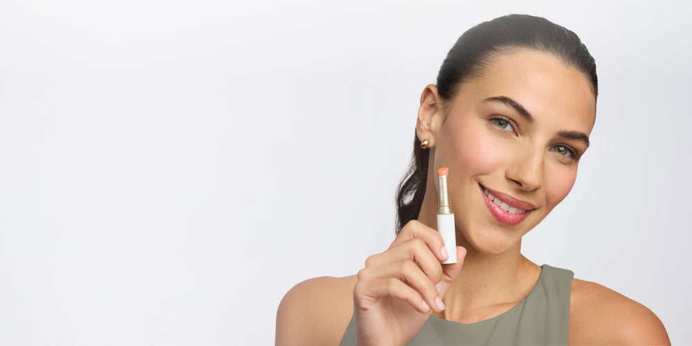 Frau mit glatter, strahlender Haut hält einen Lippenpflegestift von jane iredale in der Hand und lächelt freundlich in die Kamera vor grauem Hintergrund.