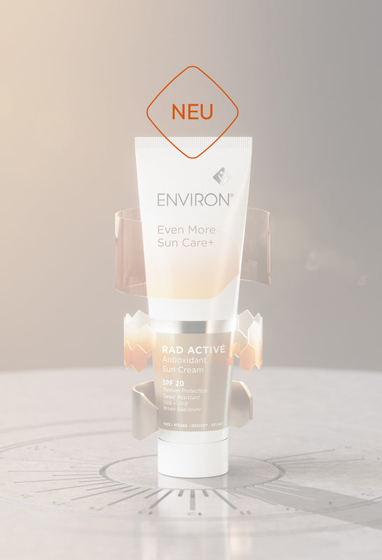 Weiße Tube der Sonnencreme „Environ RAD Activé Antioxidant Sun Cream SPF 20“ steht auf einem runden, steinernen Podest vor warm-beigem Hintergrund; goldene, grafische Elemente umgeben das Produkt, daneben der Schriftzug „NEU“