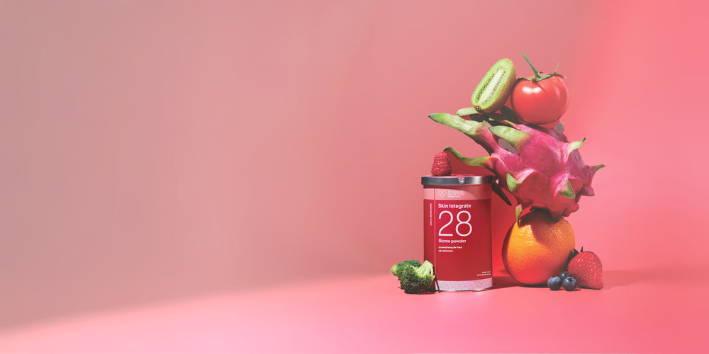 Rote Produktdose von ADVNACED NUTRITION PROGRAMME mit der Aufschrift ‚Skin Integrate 28 Biome powder‘ neben frischem Obst und Gemüse wie Kiwi, Tomate, Drachenfrucht, Orange, Himbeere, Blaubeeren, Erdbeere und Brokkoli vor rotem Hintergrund.