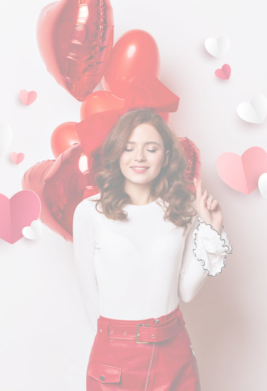 Junge Frau mit geschlossenen Augen und einem glücklichen Gesichtsausdruck steht vor einem hellen Hintergrund mit roten Herzluftballons und Papierherzen. Sie trägt ein weißes Oberteil mit Volantärmeln und einen roten Rock. Die Szene vermittelt eine romantische, verspielte Stimmung – perfekt für den Valentinstag.